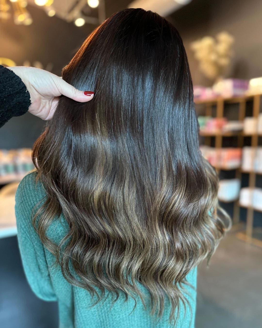 A person with long, wavy brown hair with subtle highlights is shown from behind. A hand with red nail polish is holding a section of the hair. Shelves with various items are visible in the blurred background.