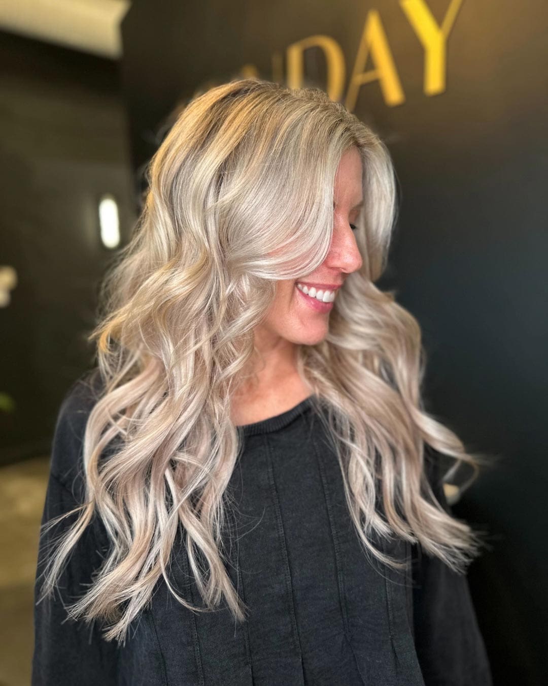 A person with long, wavy platinum blonde hair, wearing a dark top, smiles while looking to the side in a dimly lit room. The background features blurred black and yellow elements.