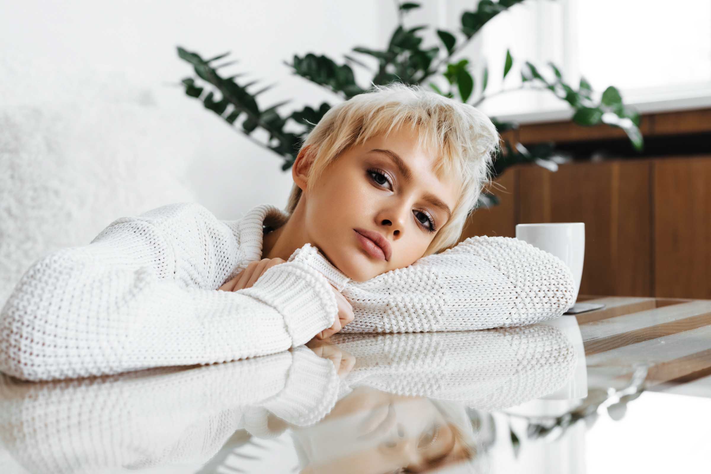 A person with short blonde hair wearing a white sweater rests their head on a table. Nearby, a white mug adds to the serene setting, while green plants provide a tranquil backdrop. The scene feels calm and reflective.