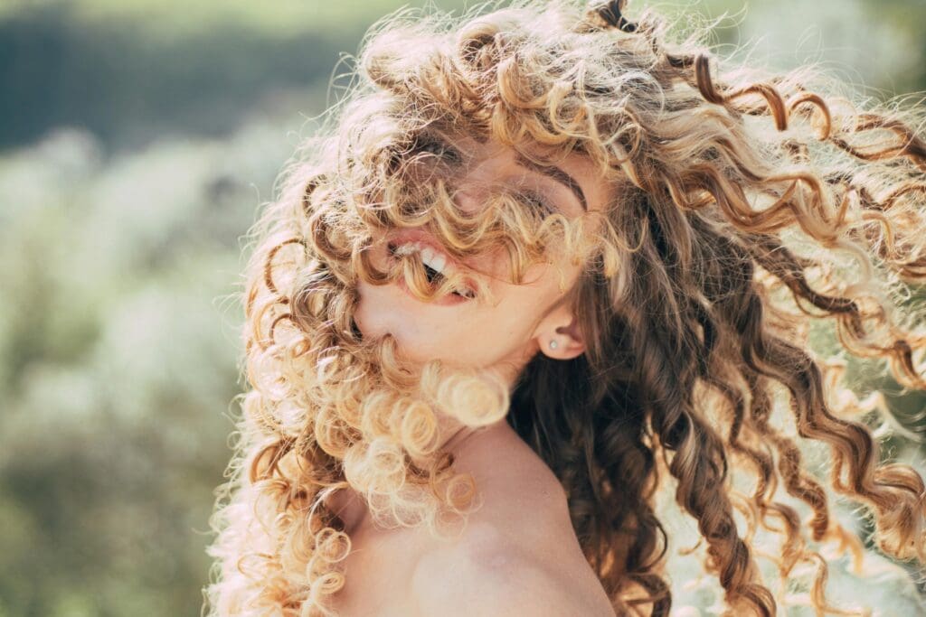 A radiant person with curly blonde hair joyfully swaying in the breeze creates a playful and lively atmosphere against a blurred green backdrop.