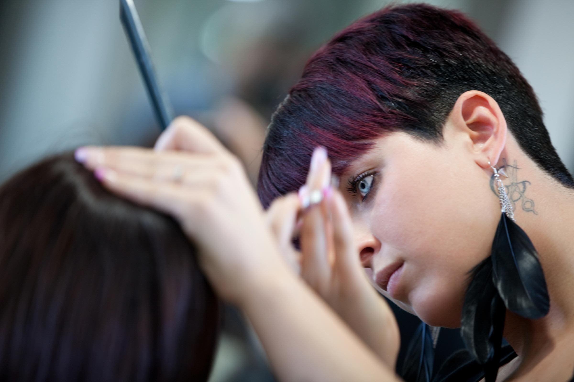 A hairstylist with short, dark hair focuses intently on delivering precision haircuts, skillfully wielding scissors and a comb. Her feather earrings sway gently as she works, and the salon setting buzzes softly around her.