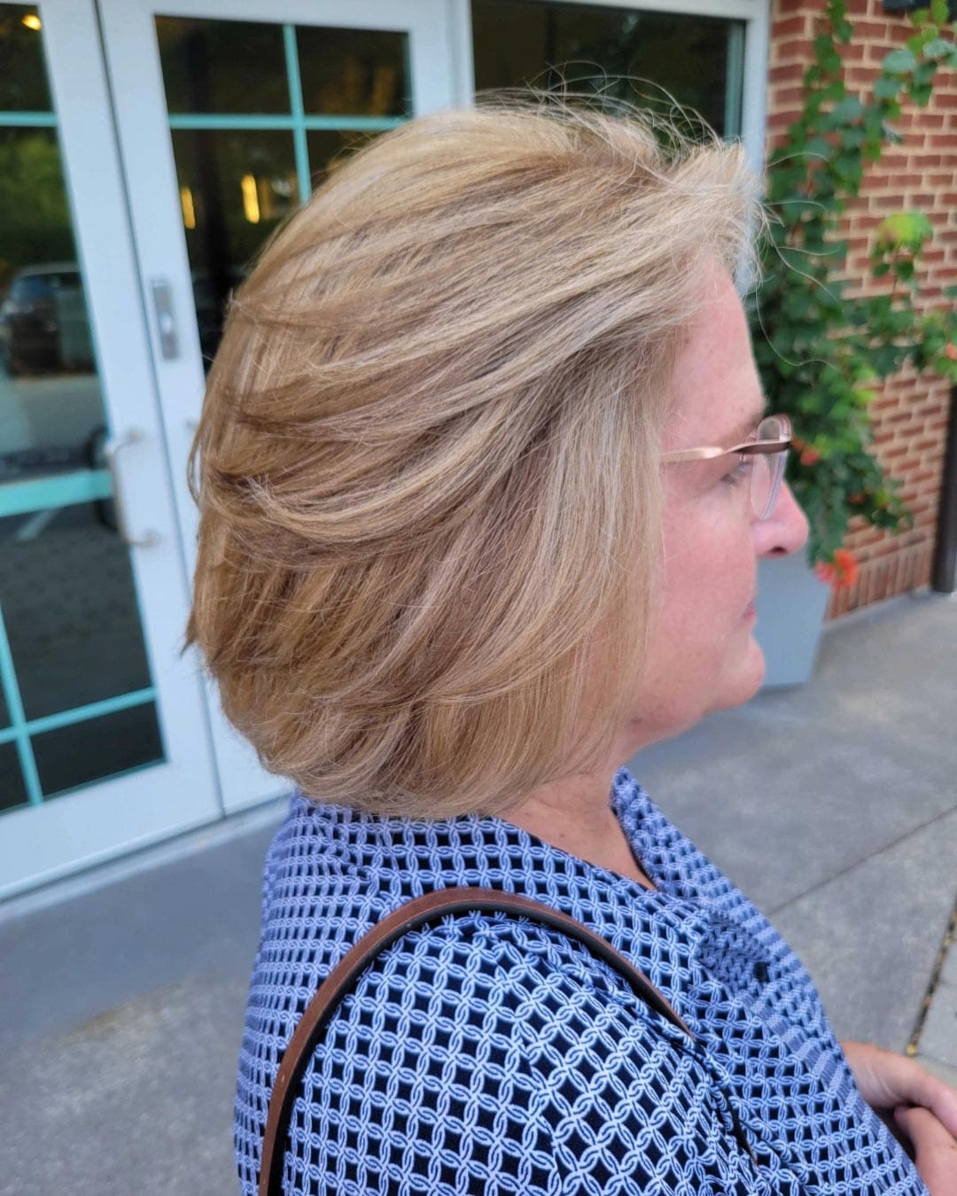 A woman with layered blonde hair and glasses stands outside, wearing a blue patterned blouse. A building with glass doors and a brick wall is visible in the background.