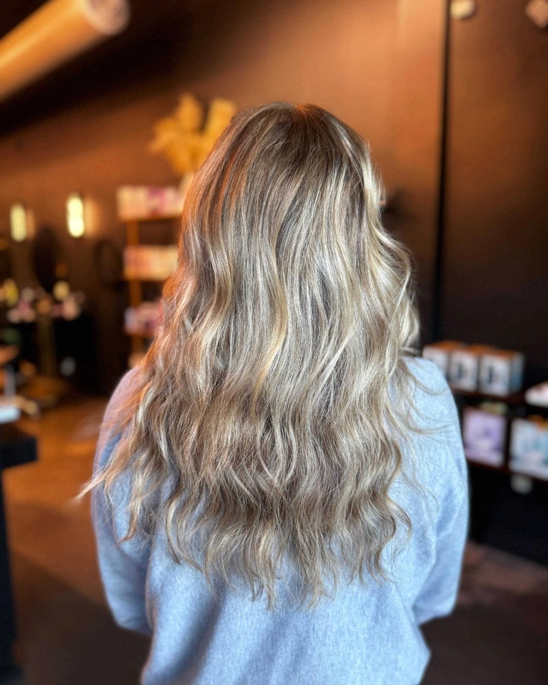 A person with long, wavy blonde hair stands with their back to the camera inside a dimly lit room. They're wearing a gray shirt, and the background shows blurred shelves with various items.