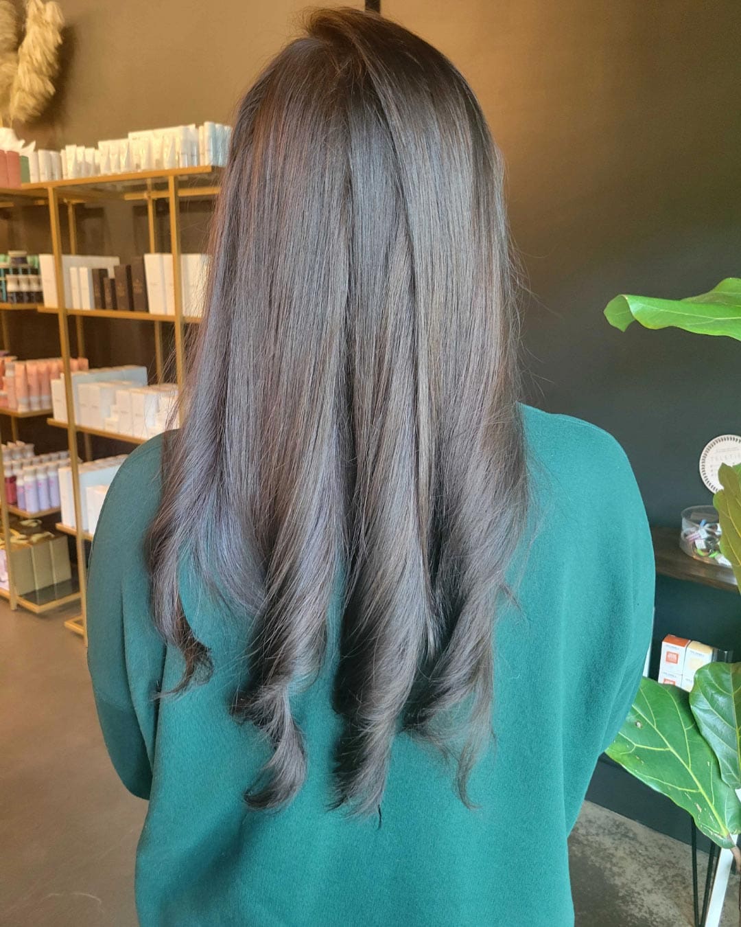 A person with long, wavy dark brown hair is seen from the back, standing in a salon with shelves of products and a green plant nearby. They are wearing a dark green top.