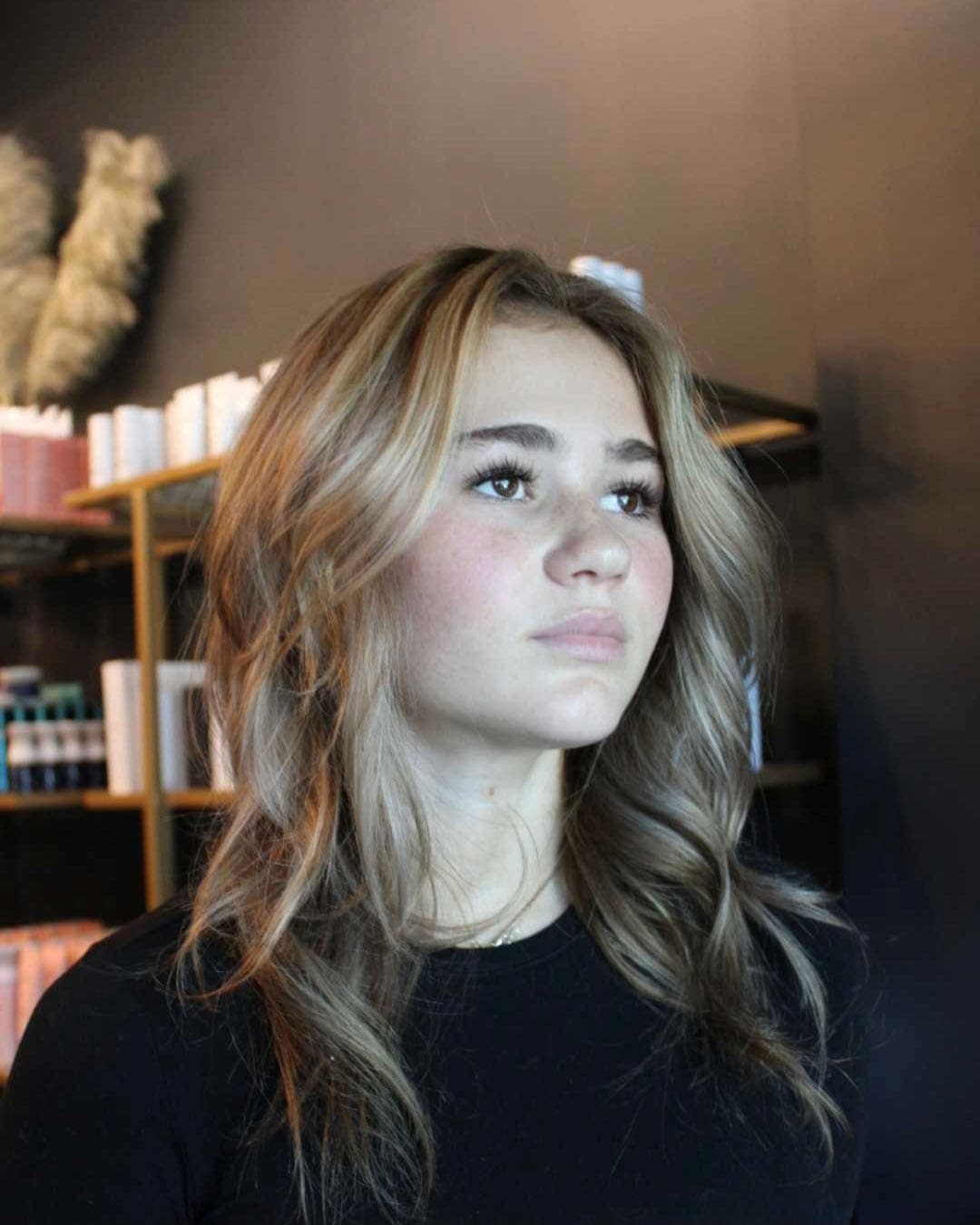 A young woman with wavy, light brown hair and a black top gazes upward. Shelves with books and decorative items are in the background. The setting appears to be indoors, possibly in a salon or a home.