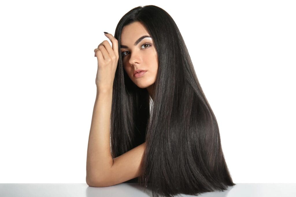 A woman with long, sleek black hair sits against a white background. She rests her chin on her hand, looking directly at the camera with a calm expression. Her glossy hair, a testament to effective hair treatments, flows over her shoulders and down the table.