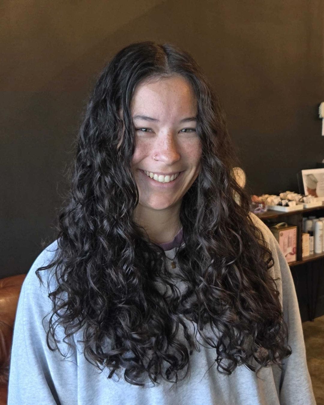 A person with long, dark, curly hair smiles at the camera. They are wearing a light-colored top. The background includes a dark wall and shelves with various items.