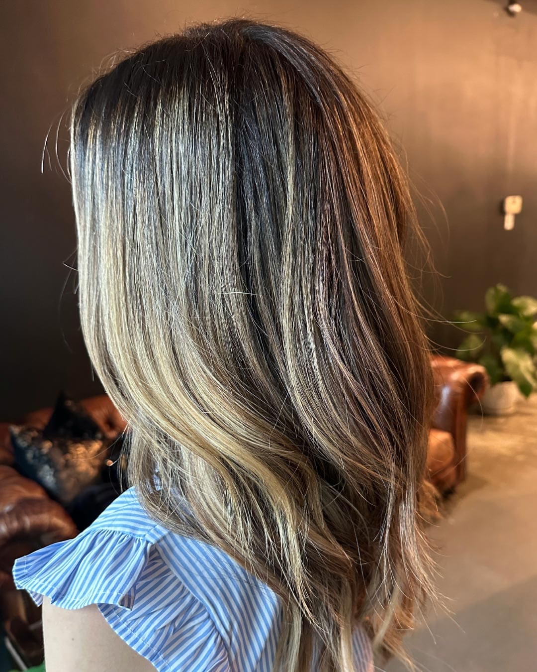 A woman with long, wavy, highlighted blonde hair is seen from the side. She is wearing a blue and white striped shirt with ruffled sleeves. The background features a brown leather chair and a plant against dark walls.