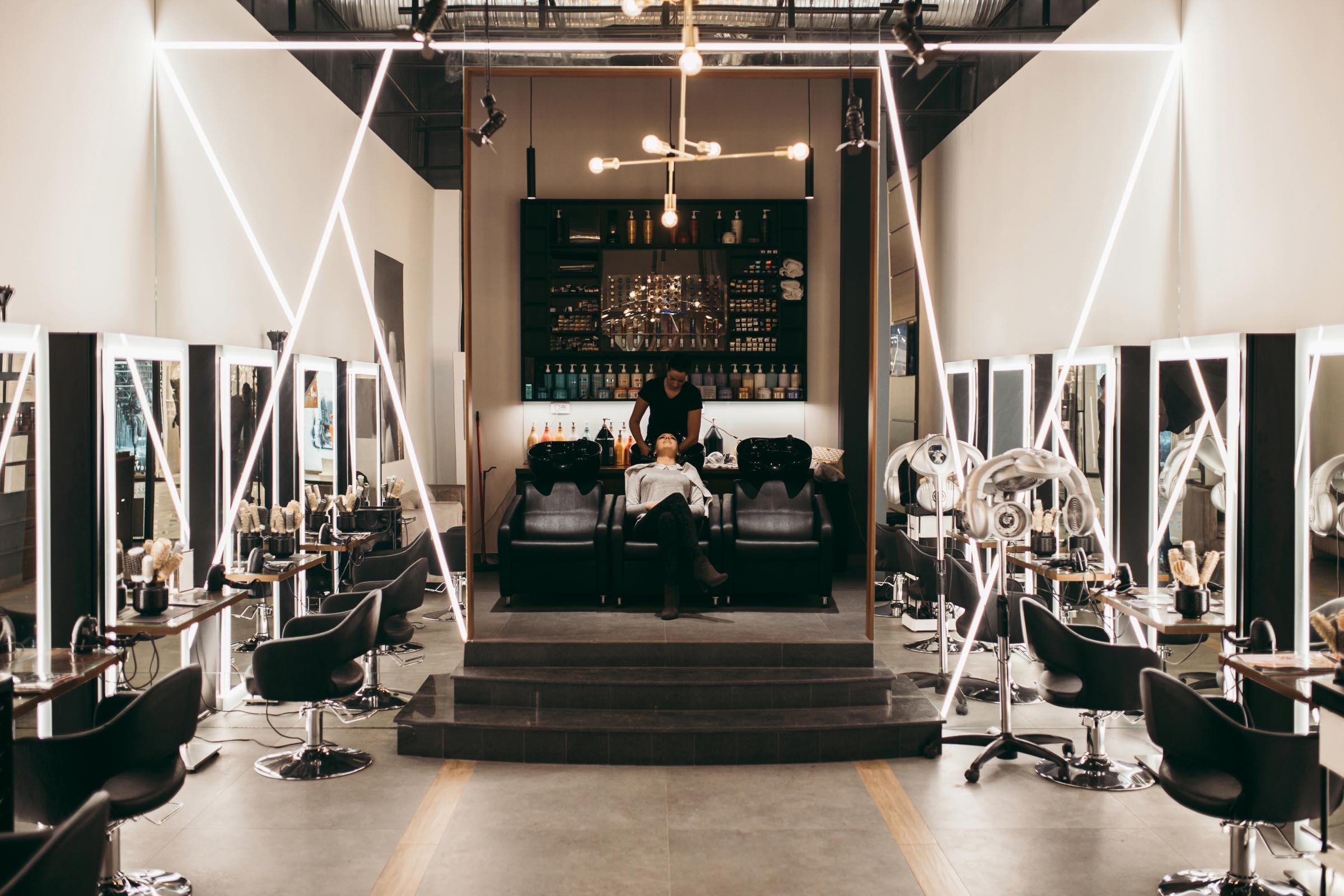 A modern, sleek hair salon in Cary, NC features stylish black chairs and symmetrical lighting. At the center, a person enjoys a relaxing hair wash as mirrors and shelves with premium products line both sides. In the background, a bar-like counter showcases an array of bottles.