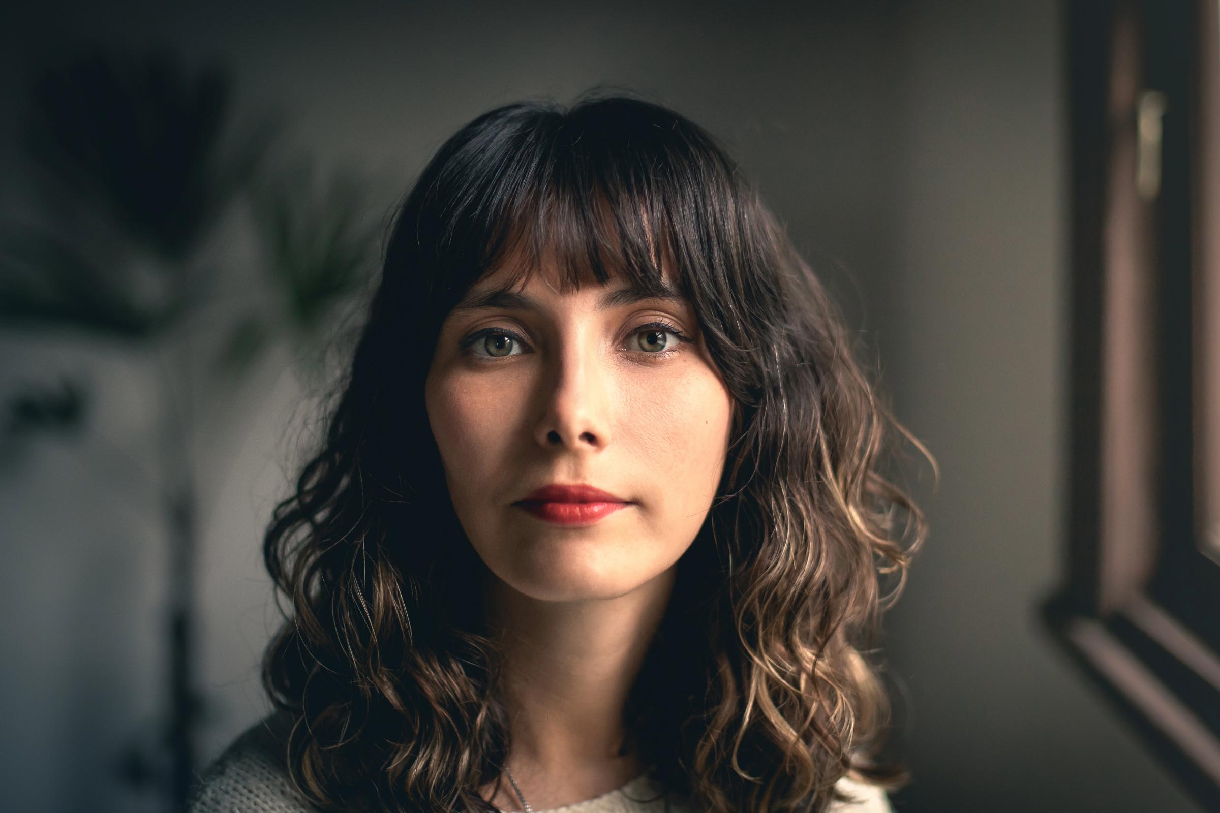 A woman with wavy brown hair and classic types of bangs, wearing a light-colored sweater, stands indoors near a window. She gazes directly at the camera with a neutral expression. The background is softly blurred, suggesting a cozy home setting.