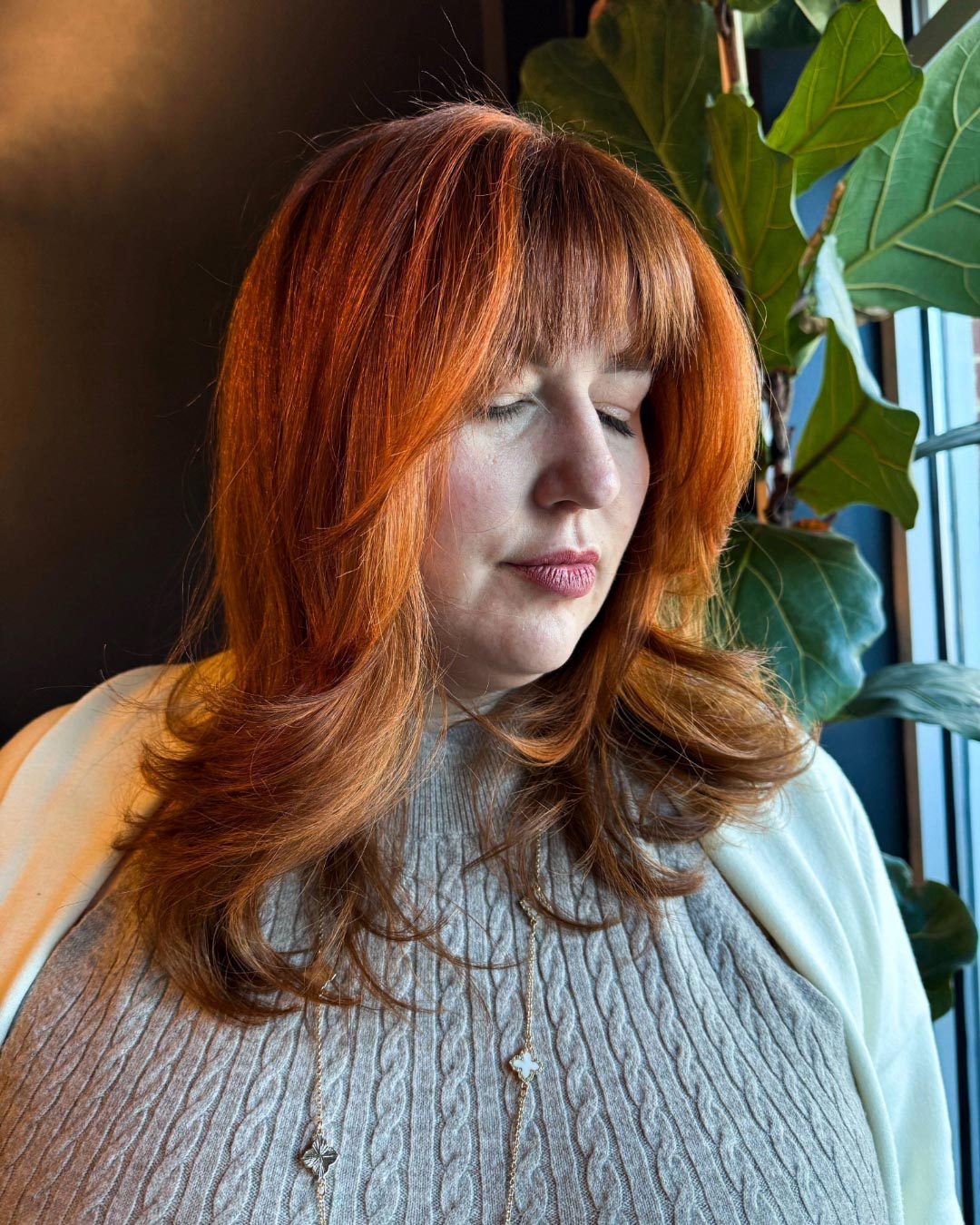 A person with long red hair and bangs is standing near a window. They are wearing a gray sweater and a light jacket. Their eyes are closed, and they are surrounded by greenery. A warm light casts a cozy glow.