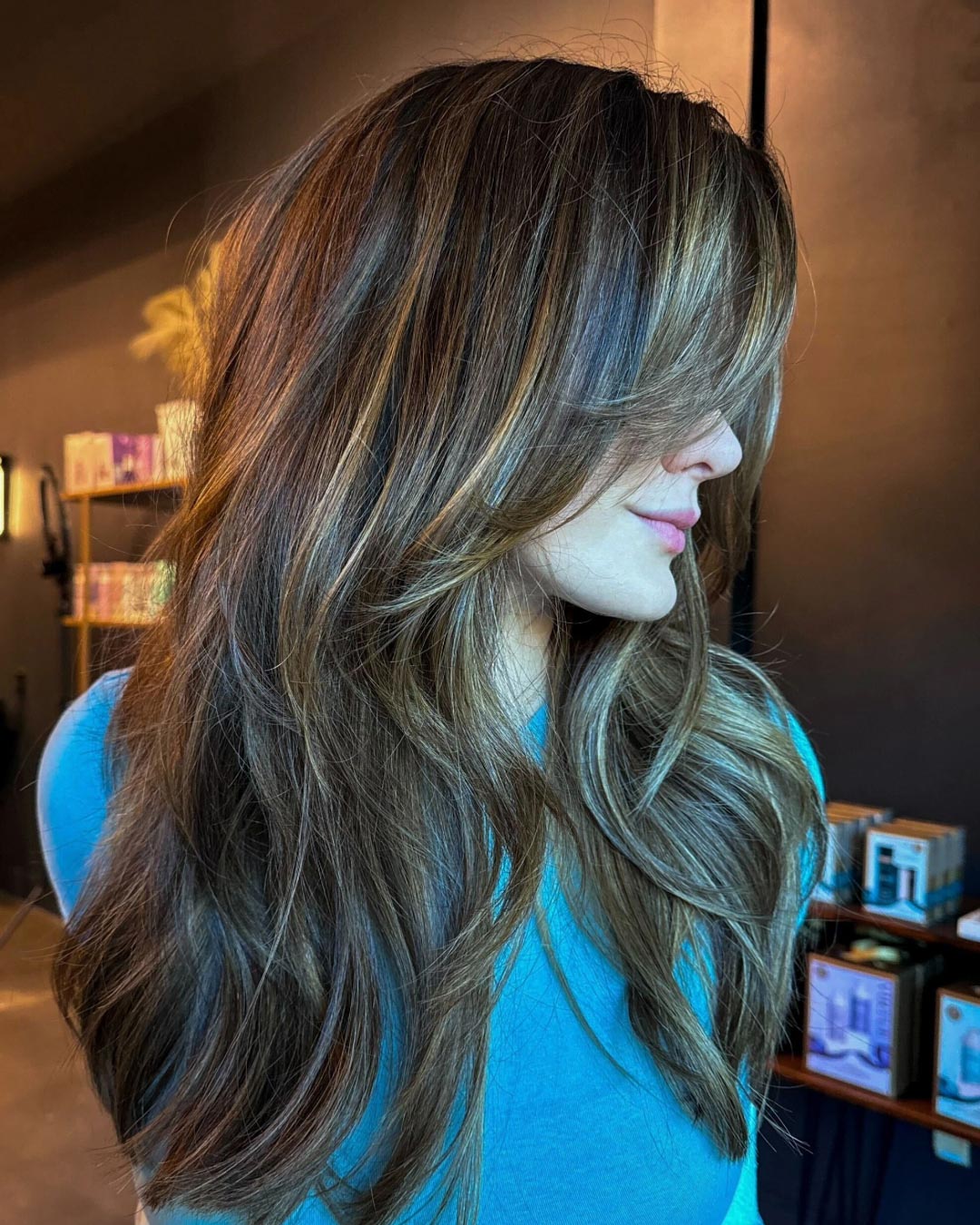 Woman with long, layered brunette hair with blonde highlights, wearing a blue top, looking down. The background shows a dark wall and shelves with various colorful items.