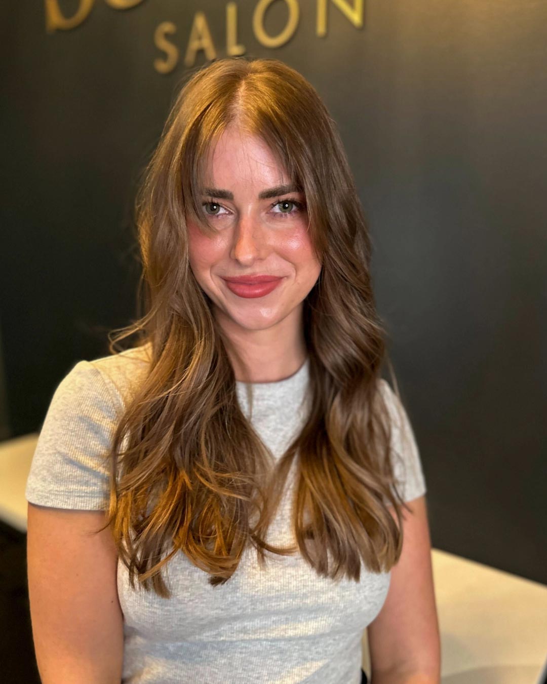 A person with long, wavy brown hair and bangs smiles while wearing a light gray top. They are standing indoors with a dark salon backdrop.