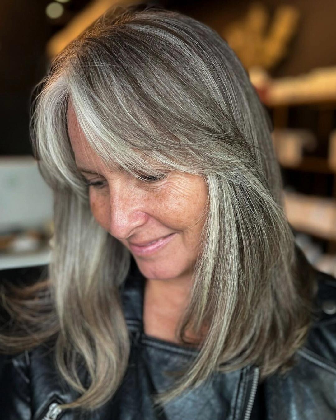 A woman with long, silver-gray hair and side-swept bangs looks down with a slight smile. She is wearing a black leather jacket, and the background is softly blurred, giving a warm ambiance.