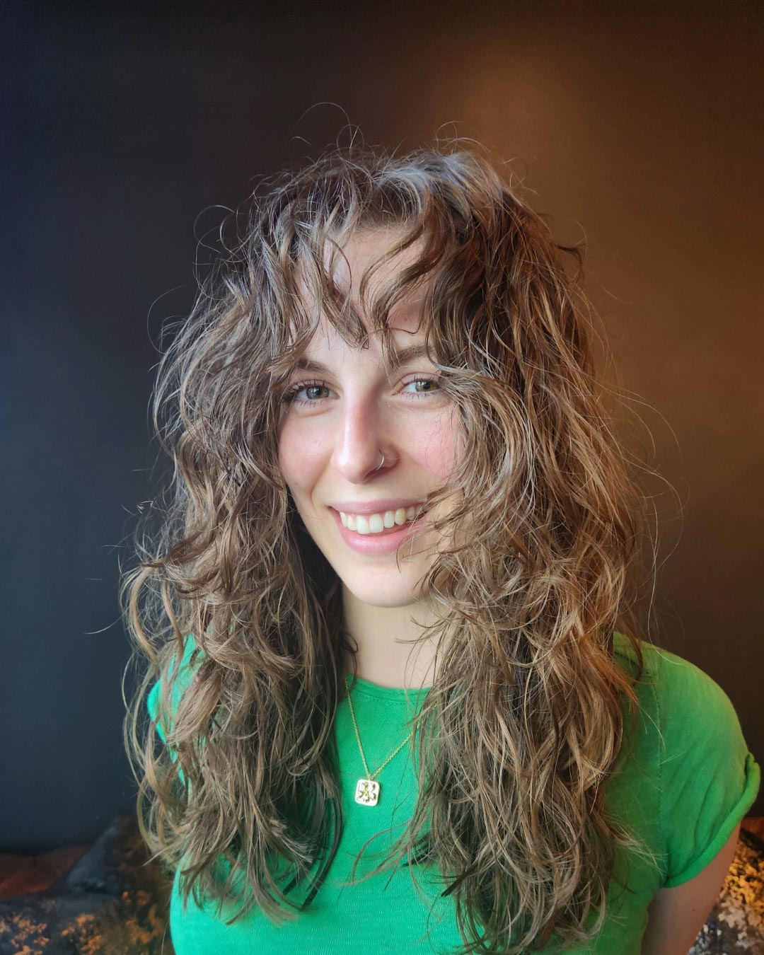 A person with wavy, light brown hair and a nose ring smiles at the camera. They are wearing a green shirt and a necklace with a square pendant. The background is a plain, dark color.
