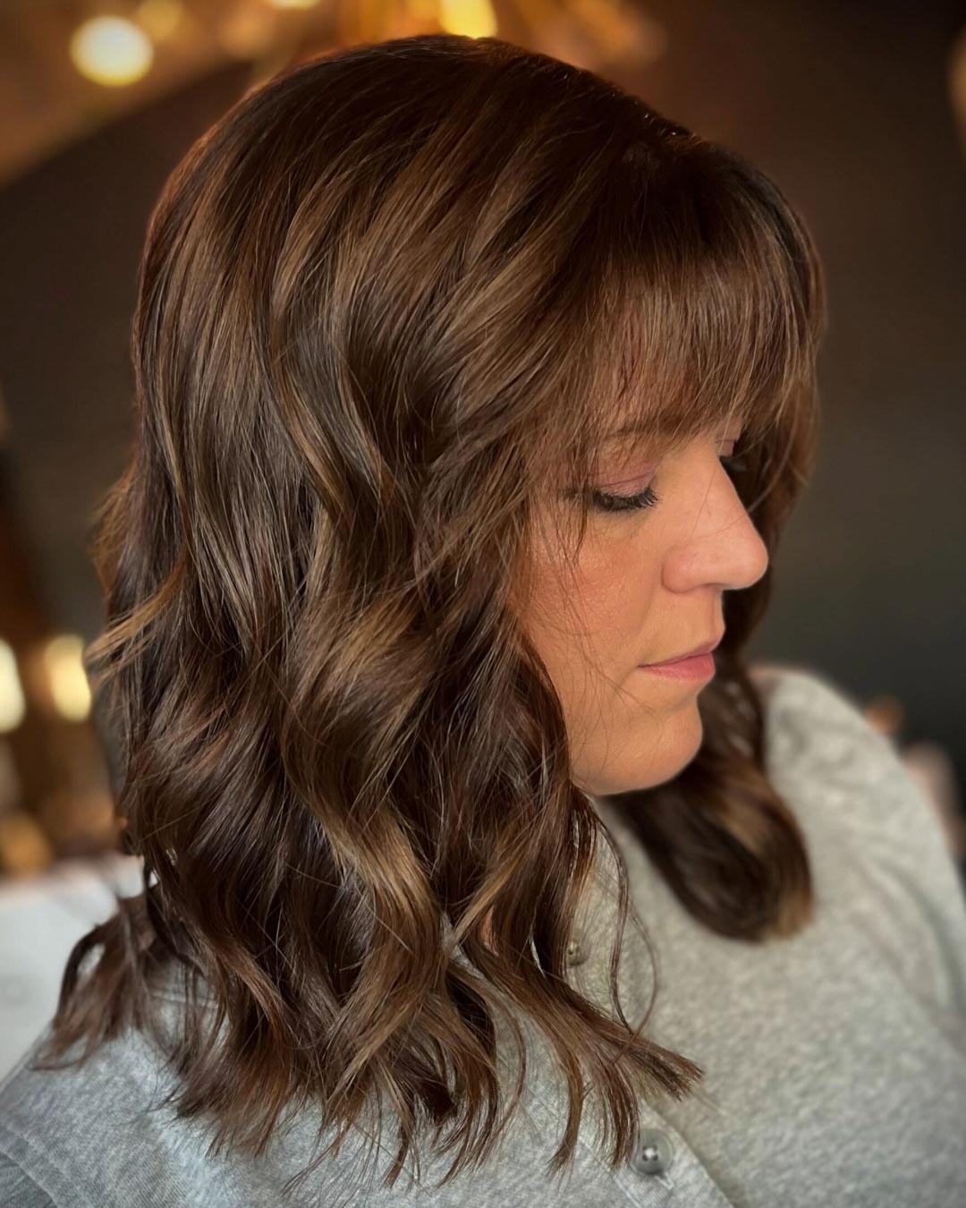 A person with medium-length, wavy brown hair and bangs looks down, wearing a gray top. The background is softly blurred, highlighting the hairstyle.