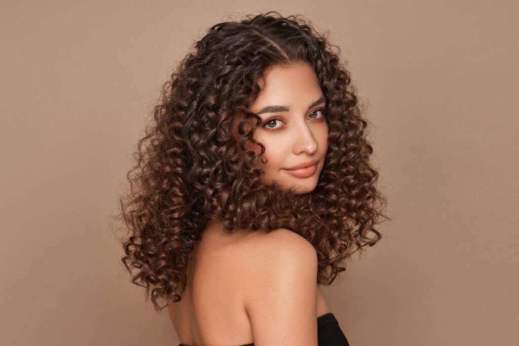 Woman with long, dark, curly hair and light skin poses in front of a beige background, looking over her shoulder with a soft smile. She wears a black, off-shoulder top—her voluminous locks styled to perfection by a curly hair salon.