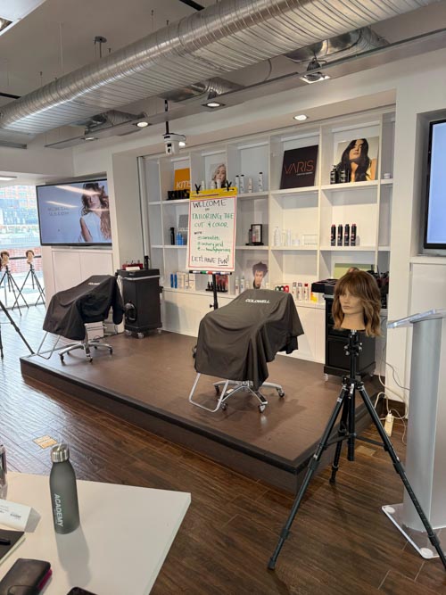 A modern salon setup with two styling chairs on a small stage, mannequin heads, hair products on shelves, and a welcome sign for a color-cutting event. Tripods and salon tools are placed around the room.