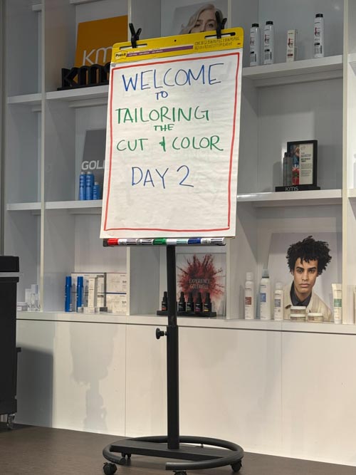 An easel holds a flip chart that reads "WELCOME TO TAILORING THE CUT & COLOR DAY 2" in a salon setting, with shelves of hair products and photos of hairstyles in the background.