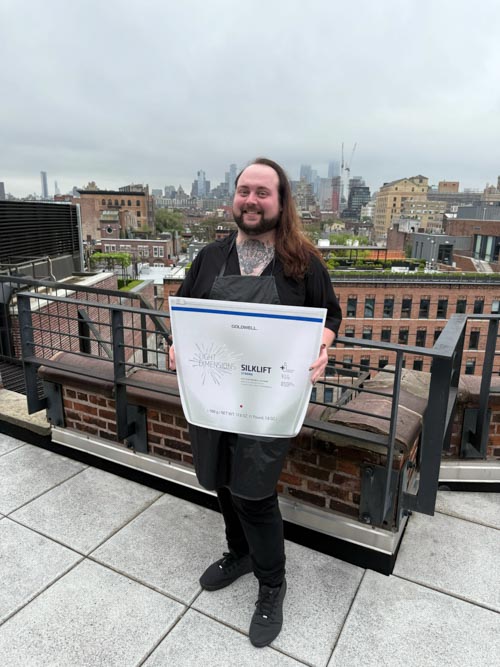 A person with long hair and tattoos smiles while holding a large container labeled “Silklift” on a rooftop with a cityscape and cloudy sky in the background.