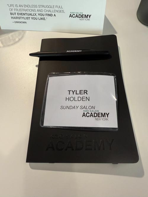 A black notebook and pen with "Kao Salon Academy" branding, a name tag reading "Tyler Holden, Sunday Salon, Kao Salon Academy New York," and a card with an inspirational quote are on a table.