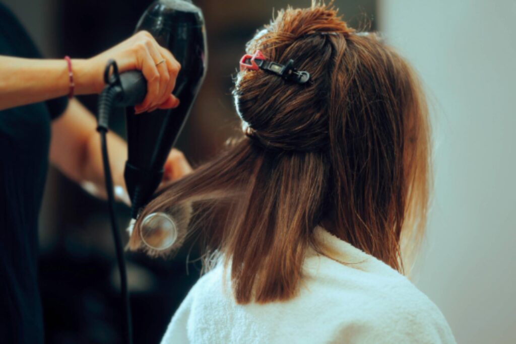 A person with shoulder-length brown hair is receiving one of the salon blowouts, with their hair partially clipped up as it’s blow-dried, while a white towel rests over their shoulders.