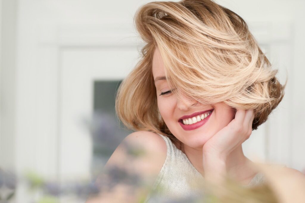 A woman with short hair and wavy blonde locks smiles with her eyes closed, resting her face in her hand. She wears a light-colored sleeveless top, and the background is softly blurred.