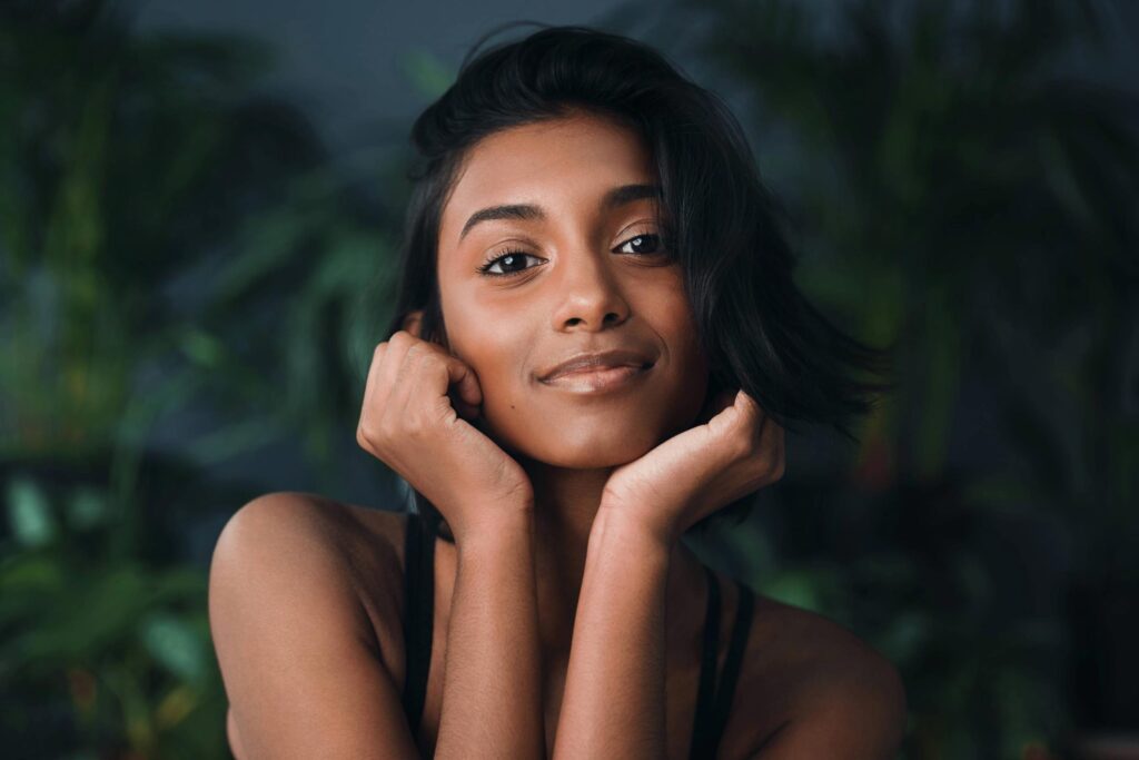 A young woman with short dark hair rests her chin on both hands, smiling softly at the camera. She is in front of a blurred green background with plants.