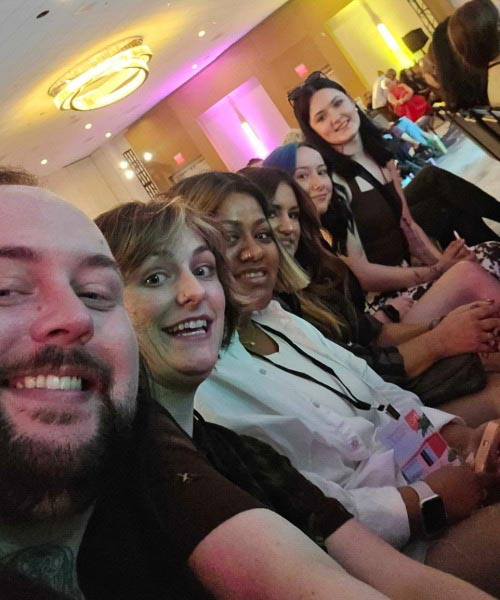 A group of six people sit closely together in a row, smiling and taking a selfie at an indoor event with colorful lighting and other attendees in the background.
