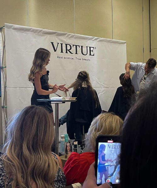 A woman demonstrates hair styling on a model's hair at a Virtue event, while another stylist works on a second model. Attendees watch, with one person recording the scene on their phone.