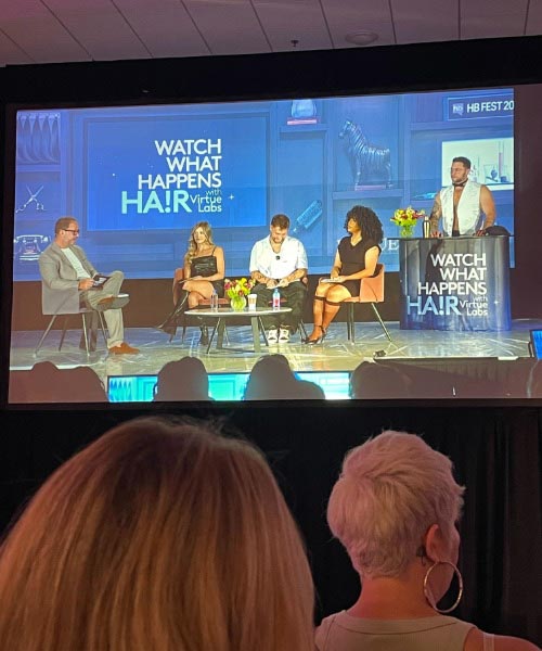 A panel of four people sits on a stage with a moderator, discussing in front of screens displaying “WATCH WHAT HAPPENS HAIR.” The audience watches attentively from the foreground.