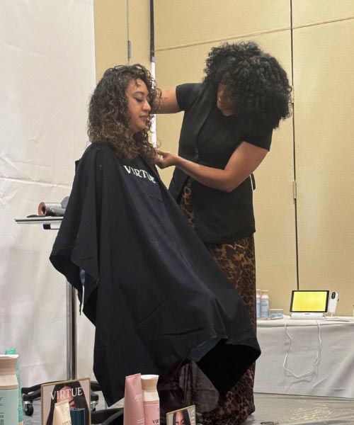A hairstylist with curly hair styles the hair of a seated woman, also with curly hair, in a salon setting. Hair products are displayed on a small table nearby. Both women appear focused on the hair styling.
