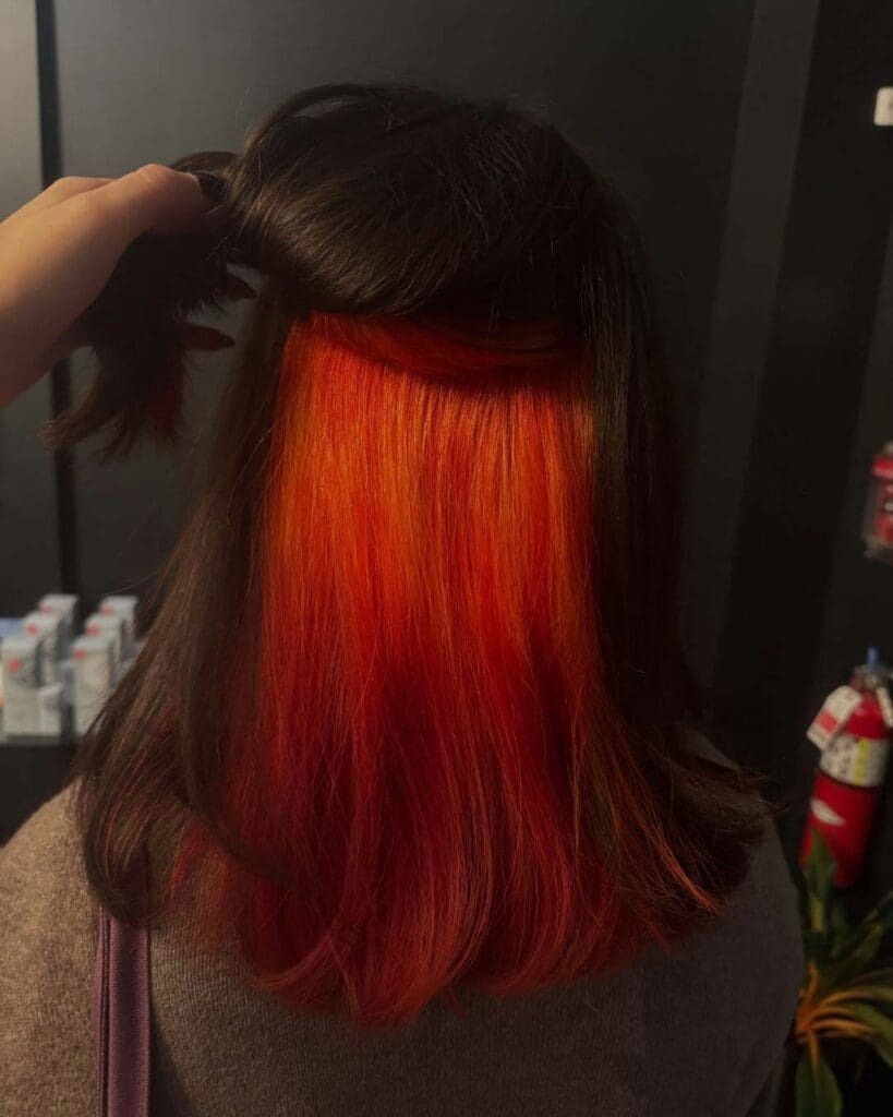 Fiery Orange Vivid Underlights with Dark Brunette Base A client holding up a sleek dark brown top layer of shoulder-length hair to reveal a bold, vibrant fiery orange and red peekaboo underlayer, expertly crafted at Sunday Salon in Raleigh.