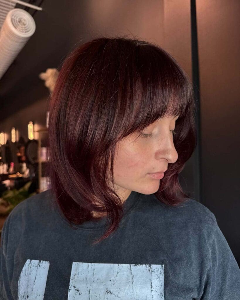 A client with a dark burgundy shoulder-length lob haircut featuring full face-framing bangs, soft texture, and rich mahogany tones, beautifully styled at Sunday Salon in Raleigh.