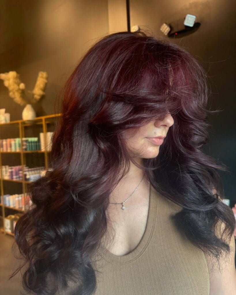 A client with long, thick hair styled in voluminous waves and sweeping curtain bangs, featuring a dimensional black cherry and deep auburn red color, styled at Sunday Salon in Raleigh.