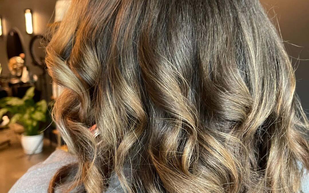 A beautifully styled client at Sunday Salon in Raleigh showcasing mid-length hair with soft, loose curls. The rich dark brunette base is seamlessly elevated with warm caramel and honey highlights, creating stunning dimension and a glossy, healthy finish that catches the light perfectly.