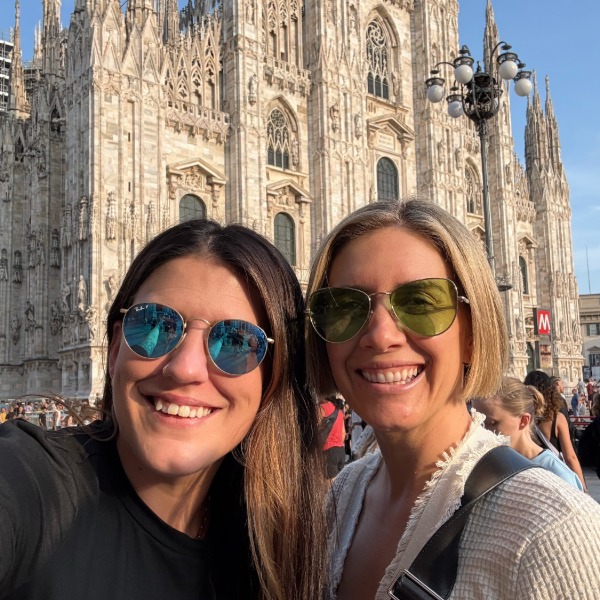 Two women wearing sunglasses smile for a selfie in front of the ornate façade of Milan Cathedral (Duomo di Milano) on a sunny day, with other people and a lamppost in the background.