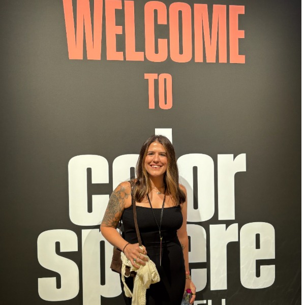 A woman with long brown hair and tattoos smiles while standing in front of a black wall with large text that reads "Welcome to Color Sphere." She is holding a drink and a cloth.