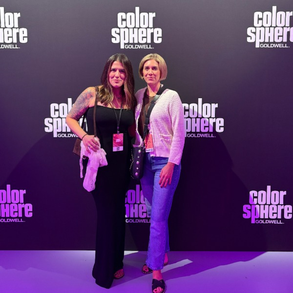 Two women pose together at an event in front of a black backdrop with the "color sphere Goldwell" logo repeated in white. One wears a black dress, the other jeans and a blazer. Both wear event badges.