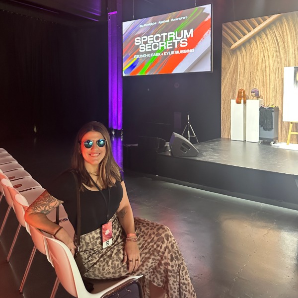 A woman wearing sunglasses and a patterned skirt sits smiling in a row of white chairs at an event. Behind her, a stage displays a colorful sign reading "Spectrum Secrets." The venue is dimly lit with purple accents.