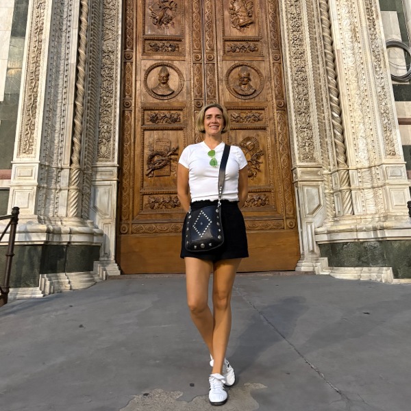 A woman in a white t-shirt, black skirt, and sneakers stands smiling in front of a large, ornate wooden door with carved details and stone columns.