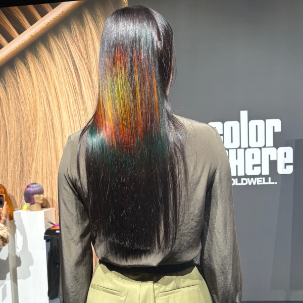 A person with long, straight black hair, featuring vibrant streaks of yellow, orange, and green on the back, stands facing away from the camera at a hair color exhibition.