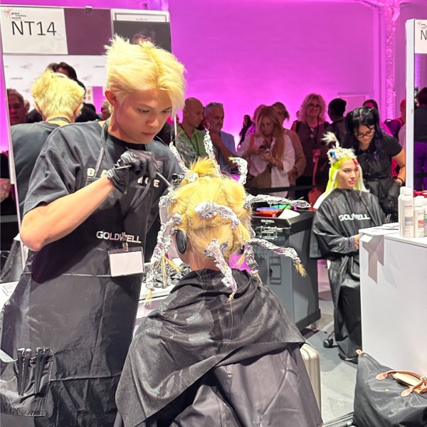A hairstylist applies hair color to a seated blonde person with foils in their hair at a salon event. Other people and stylists work and observe in the brightly lit, pink-accented room.