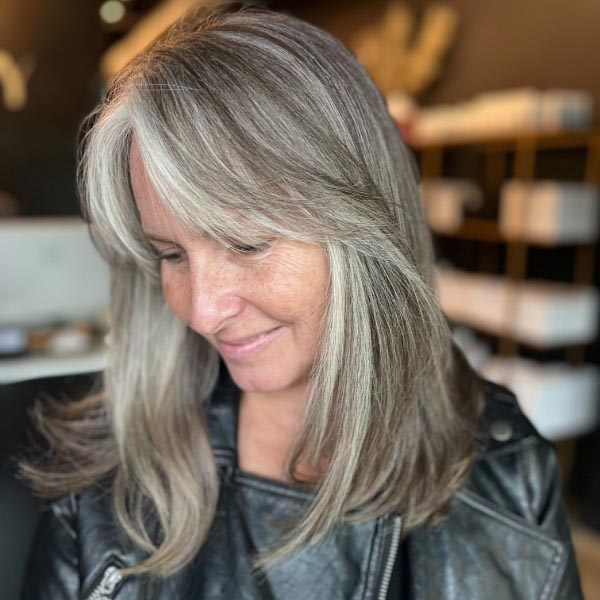 A woman with long, straight gray hair and side-swept bangs looks down and smiles gently. She is wearing a black leather jacket, and the background appears to be a modern indoor setting.