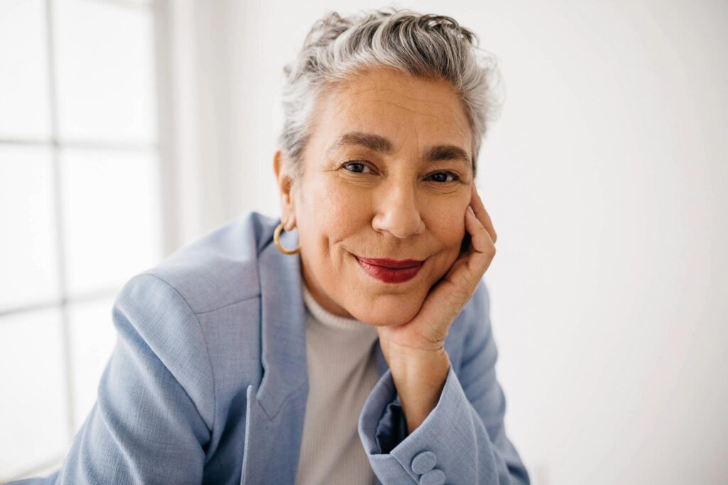 A smiling older woman with short gray hair showcases stylish grey coverage as she rests her chin on her hand. She wears a light blue blazer, white top, gold hoop earrings, and red lipstick, with a bright window in the background.