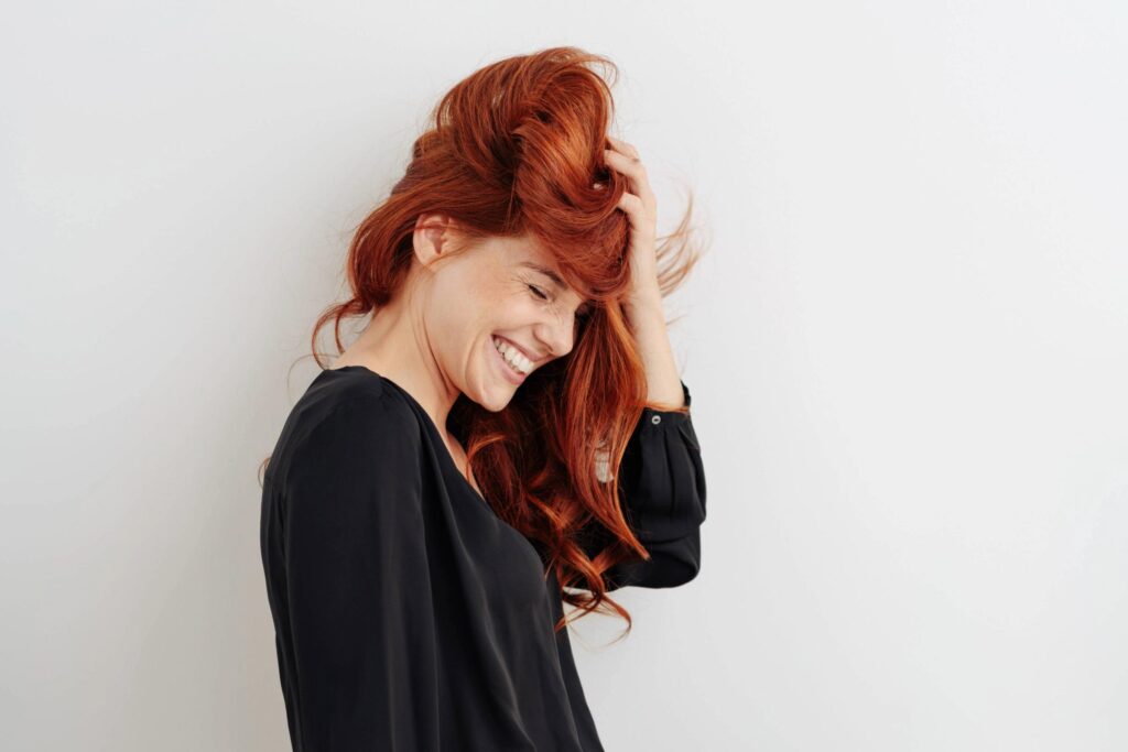 A woman with striking red hair wearing a black shirt smiles and runs her fingers through her hair while standing against a plain white background.