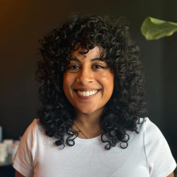 A woman with curly dark hair and a nose ring smiles at the camera, wearing a white shirt. The background is dark with a green leaf visible on the right side.