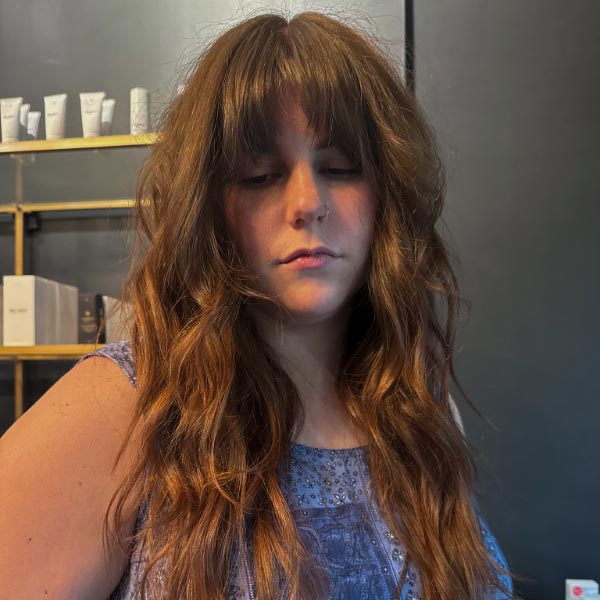 A woman with long, wavy brown hair and bangs is standing indoors, wearing a purple patterned top. Shelves with various bottles and boxes are visible in the background.