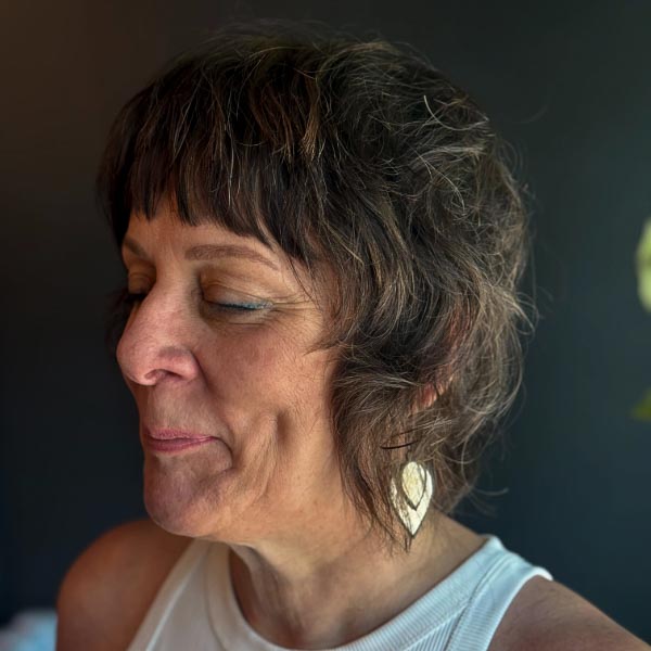 A woman with short, wavy brown hair and closed eyes wears a sleeveless white top and large, leaf-shaped earrings, set against a dark, blurred background.