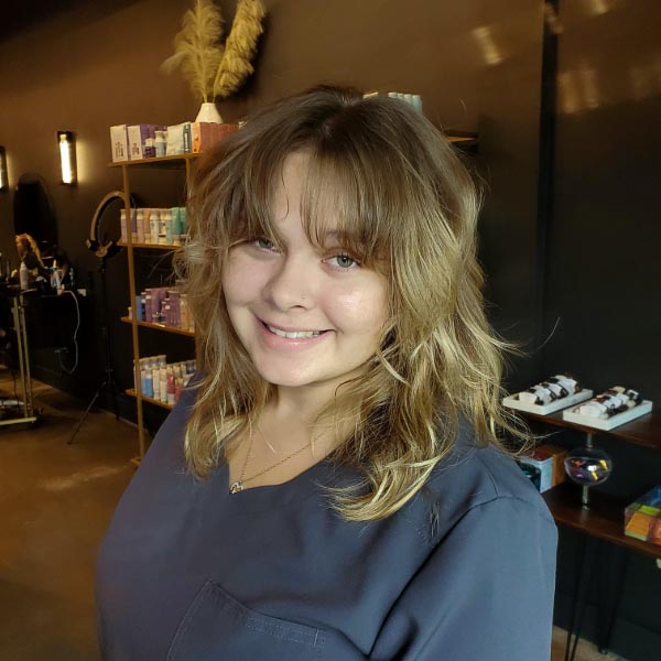 A woman with shoulder-length, wavy, light brown hair and bangs smiles in a salon with dark walls and shelves holding hair products in the background. She is wearing a dark-colored top.